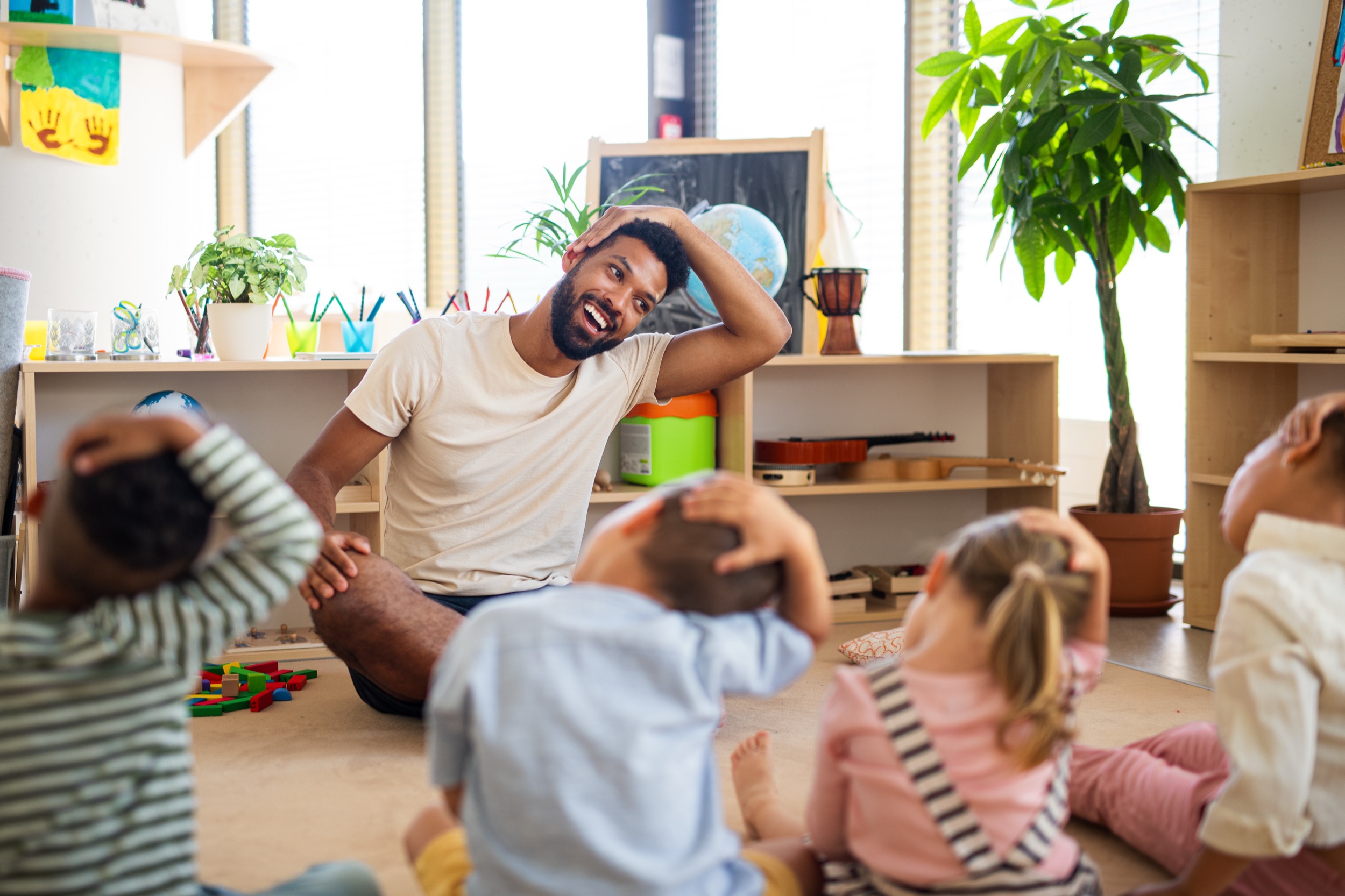 Group of small nursery school children with man teacher sitting on floor indoors in classroom, doing exercise.